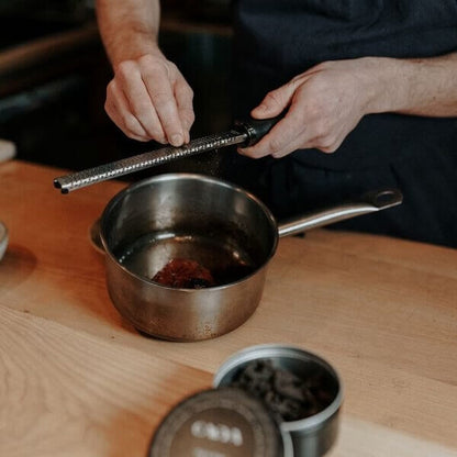 un chef utilise une râpe pour moudre du poivre long noir au dessus d'une casserole